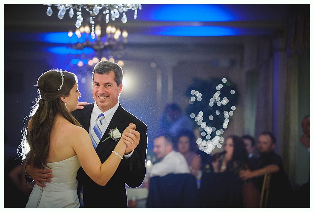 Bride with arms up, making a funny face, groom looking at his hand, cake cutting in background.