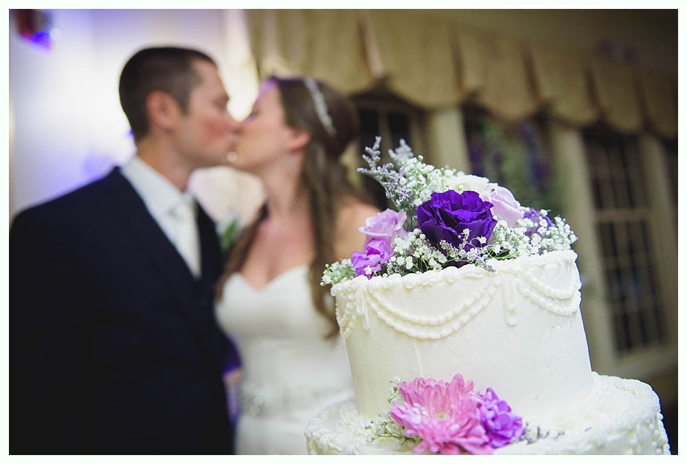 Bride with arms up, making a funny face, groom looking at his hand, cake cutting in background.