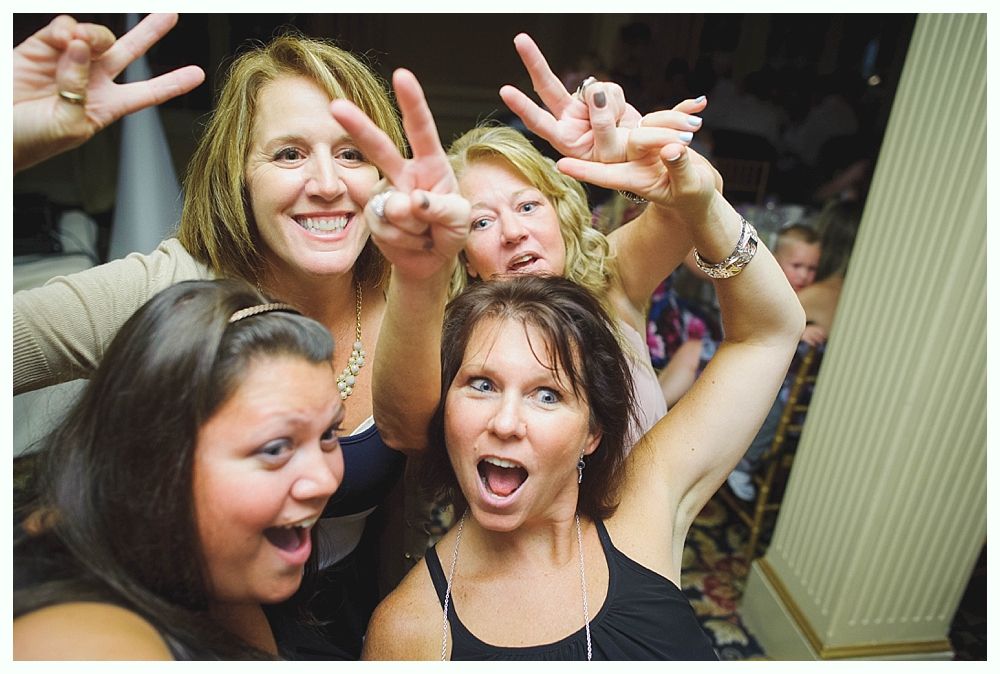 Bride with arms up, making a funny face, groom looking at his hand, cake cutting in background.