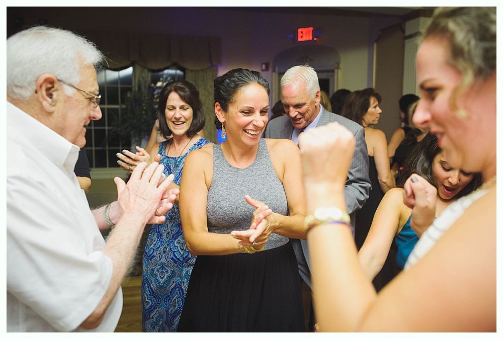 Bride with arms up, making a funny face, groom looking at his hand, cake cutting in background.