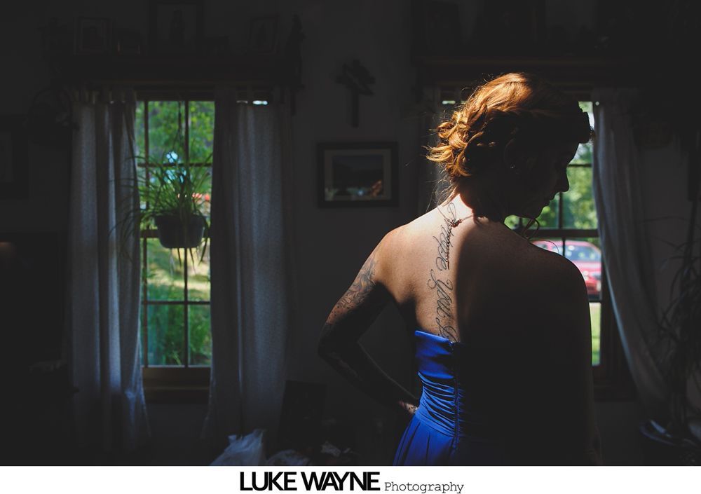 Woman with tattoo, in shadow, stands near window, light illuminating her hair and back.