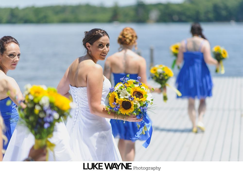 Bridesmaid in blue dress, holding sunflower bouquet, walks along a wooden dock with other bridesmaids, sunny day.
