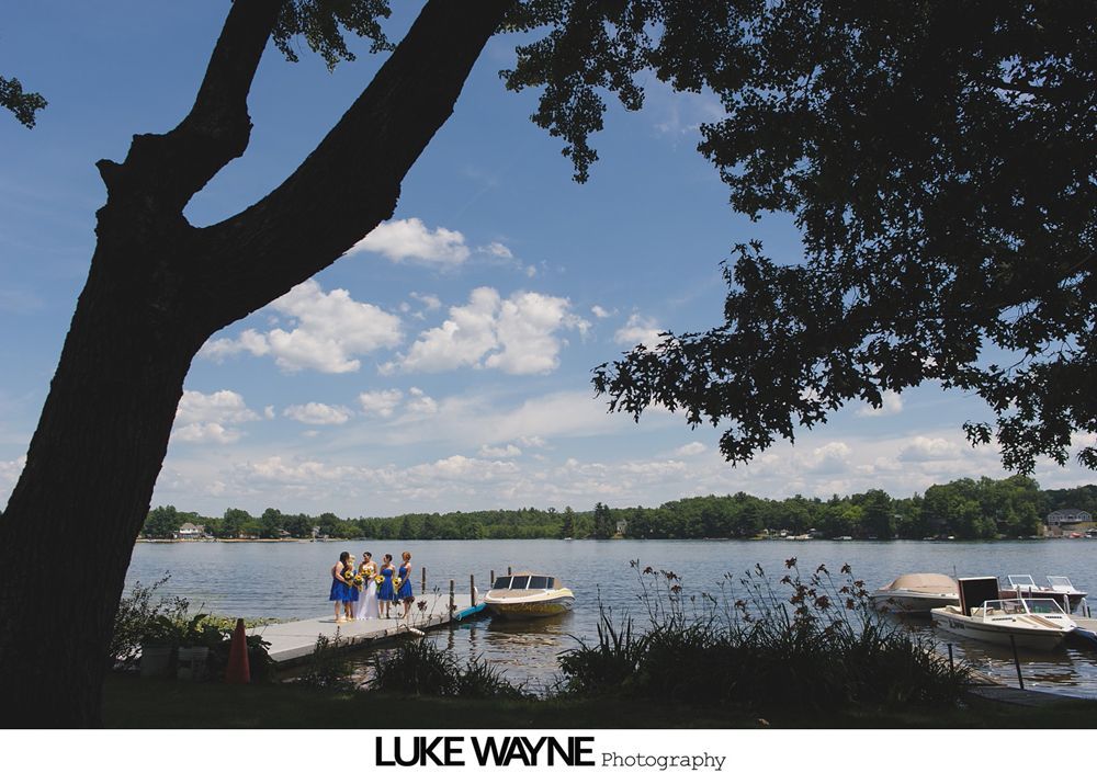 Bridesmaids in blue dresses stand on a dock by a lake, with boats and trees under a blue sky.