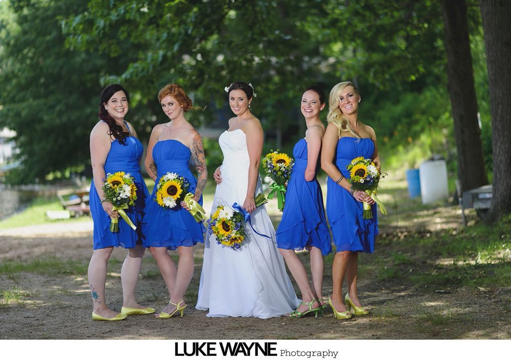 Bride and bridesmaids in blue dresses with sunflower bouquets posing outdoors.
