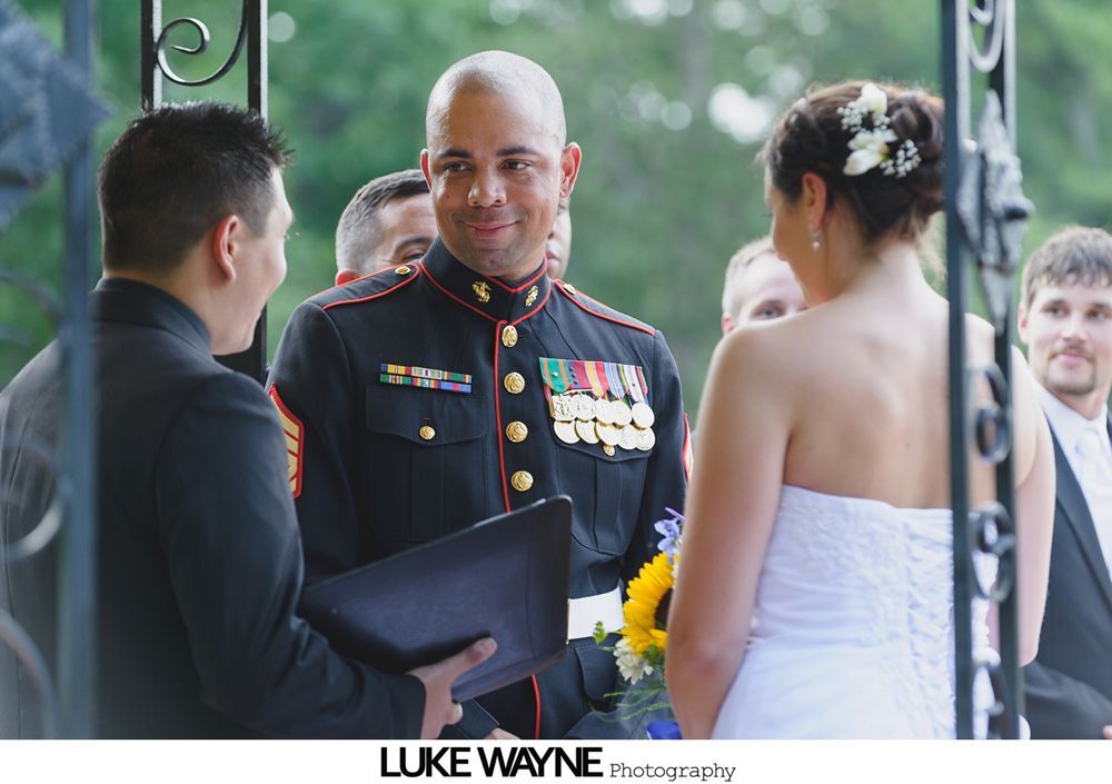 Groom in Marine uniform smiles at bride during outdoor wedding ceremony. An officiant holds a book.