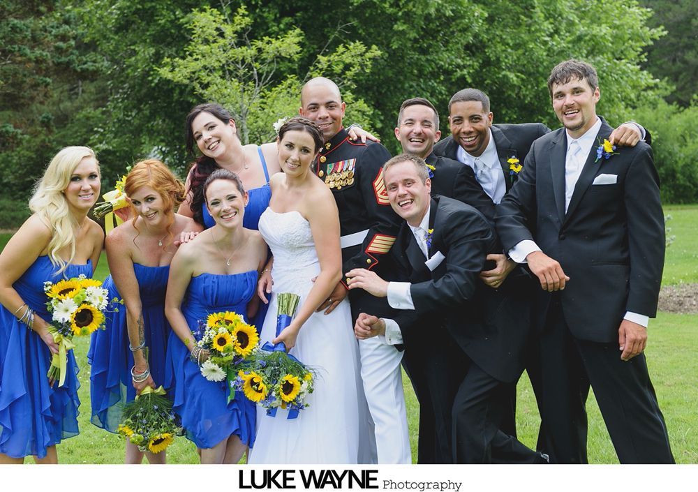 Wedding party poses outdoors; bride in white dress, bridesmaids in blue with sunflowers, groomsmen in suits.