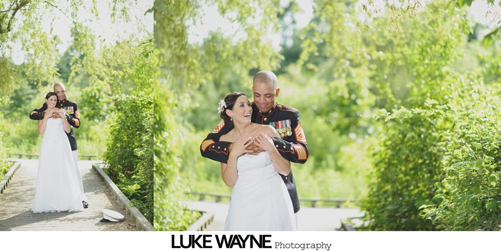 Bride and groom embrace outdoors. Man in uniform, woman in white dress, smiling. Green foliage background.