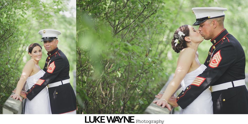 Bride and groom, in uniform, kissing on a bridge. Green foliage surrounds them.