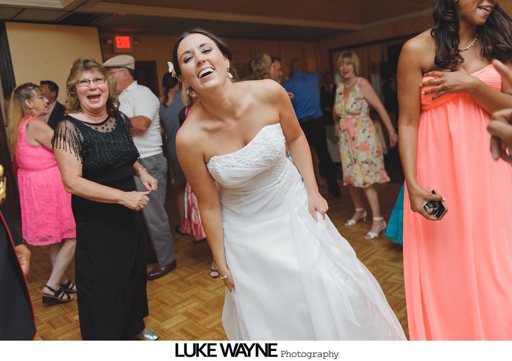 Bride laughing, dancing in white strapless gown at reception. Other guests, including one in pink, are visible.