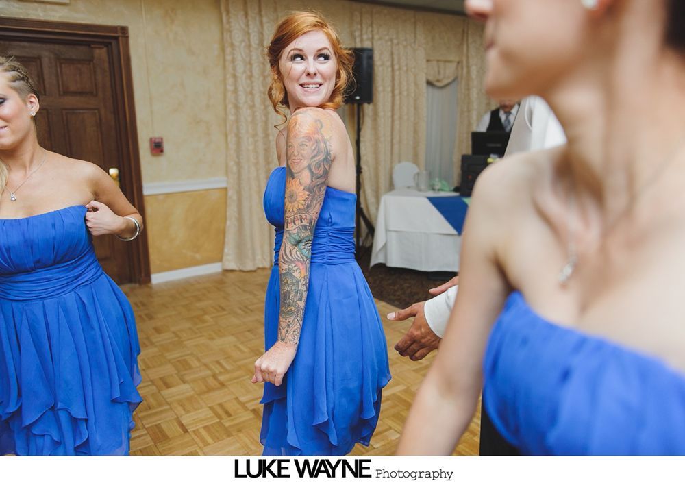 Woman with arm tattoo, in blue dress, smiles at camera on dance floor.