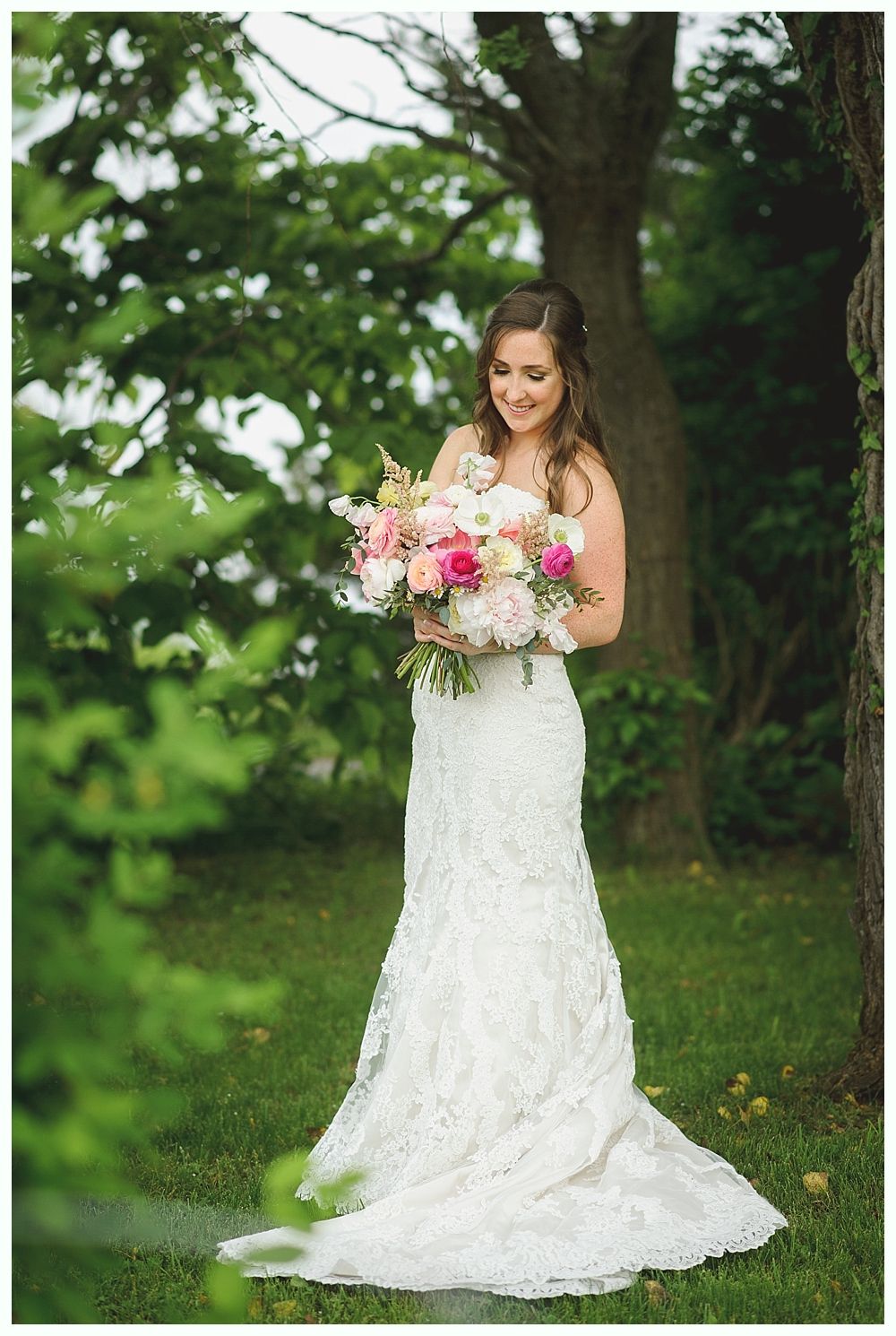 Couple embraces outdoors, framed by leaves. Groom in suit, bride in veil, near a building.