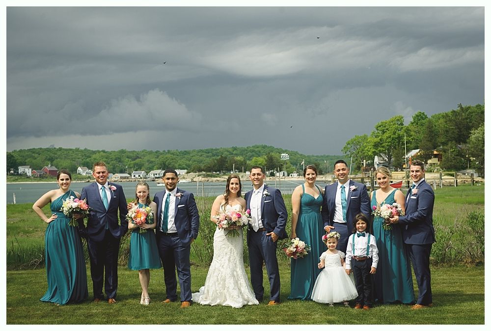 Wedding party poses in front of a red barn. Bride in white gown with bridesmaids in blue dresses, groomsmen in gray suits.
