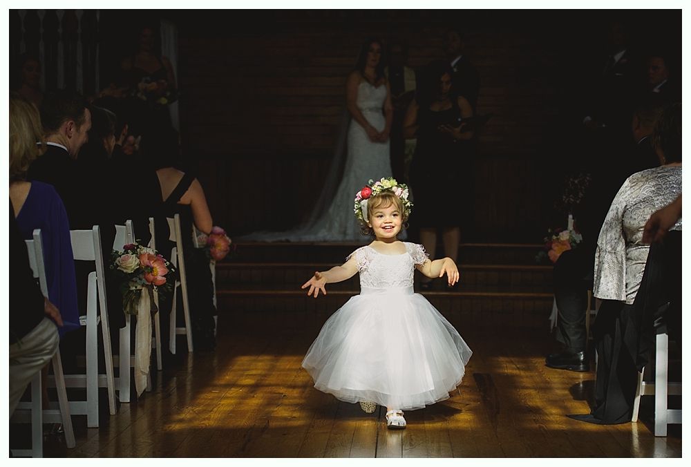 Wedding ceremony in a rustic barn with couple, guests, floral arch, and chandelier.