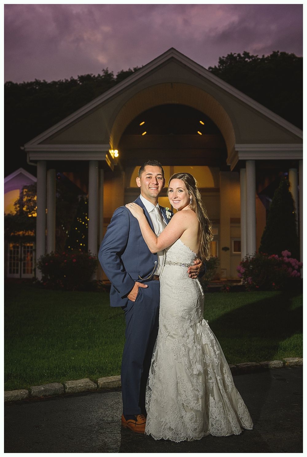 Couple in wedding attire embrace in front of a building with columns, under a cloudy sky.