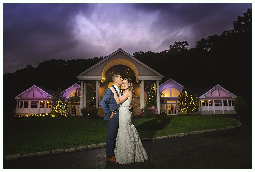 Couple embraces in front of a wedding venue at night, lit by exterior lights, dramatic sky.