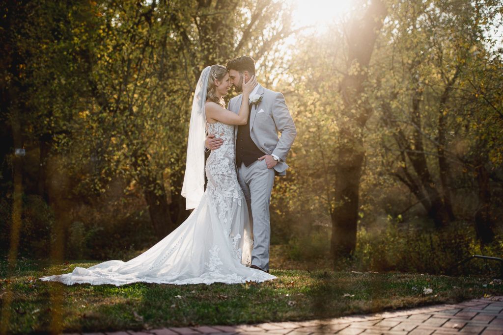 Bride and groom embrace in a sunlit outdoor setting. The bride wears a white gown, and the groom a gray suit.