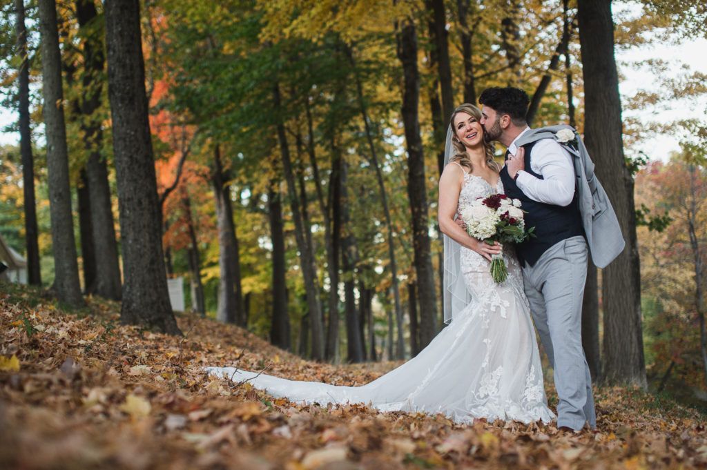 Wedding couple in a forest of fall leaves. Groom kisses bride's cheek; she smiles, holding bouquet.