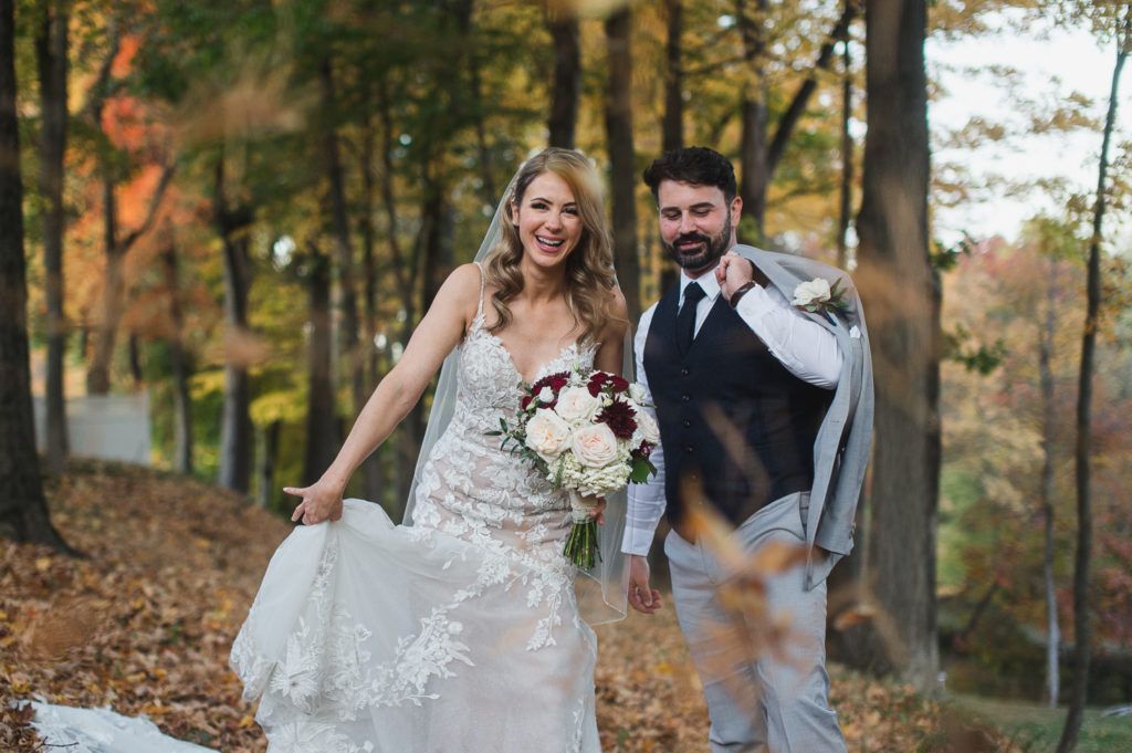 Bride and groom pose in a forest, holding bouquets, with autumn leaves falling.
