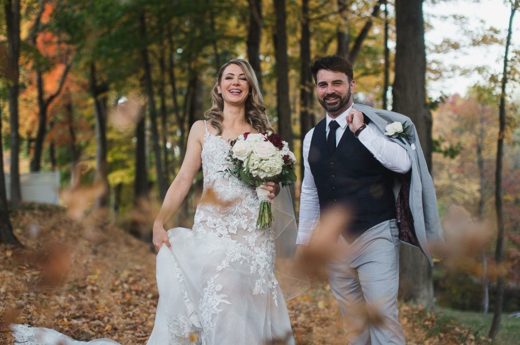 Bride and groom walking in a fall forest, holding a bouquet, leaves in the air.