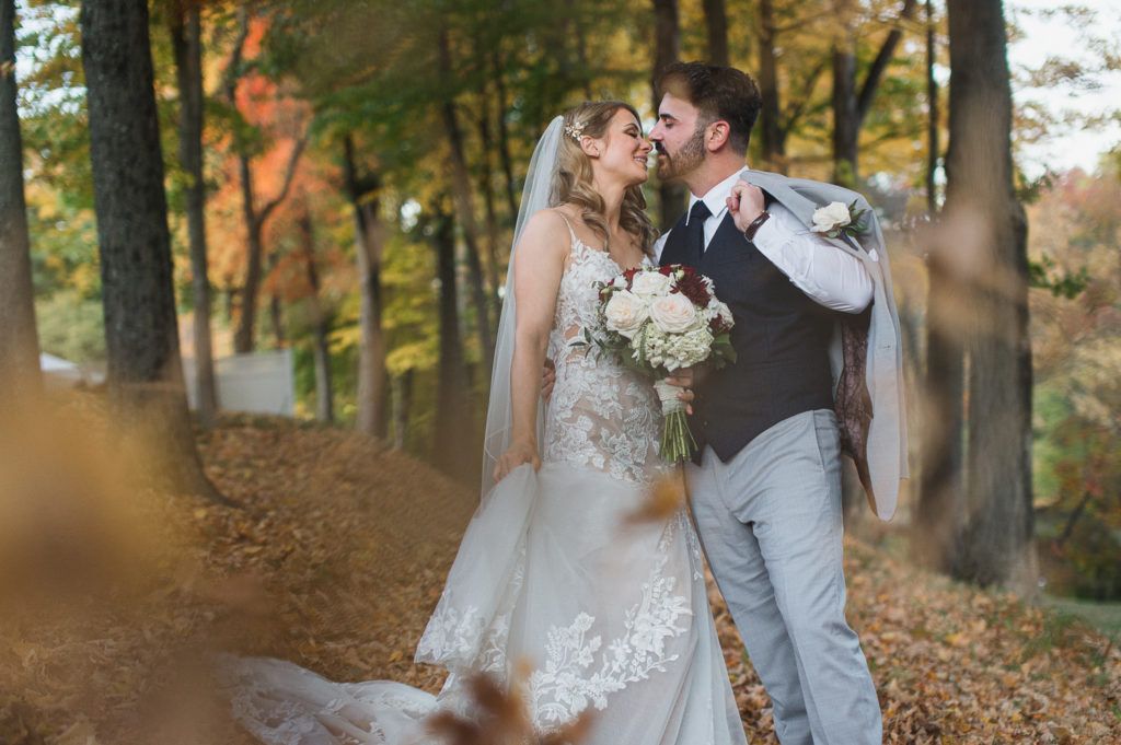 Newlyweds kissing outdoors among fall leaves; bride in white gown, groom in suit with coat draped over shoulder.
