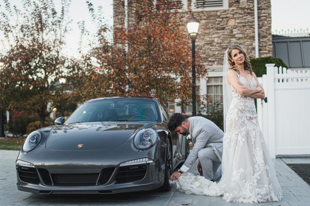 Bride in white dress watches groom adjusting dress near a grey sports car. Building and foliage in the background.
