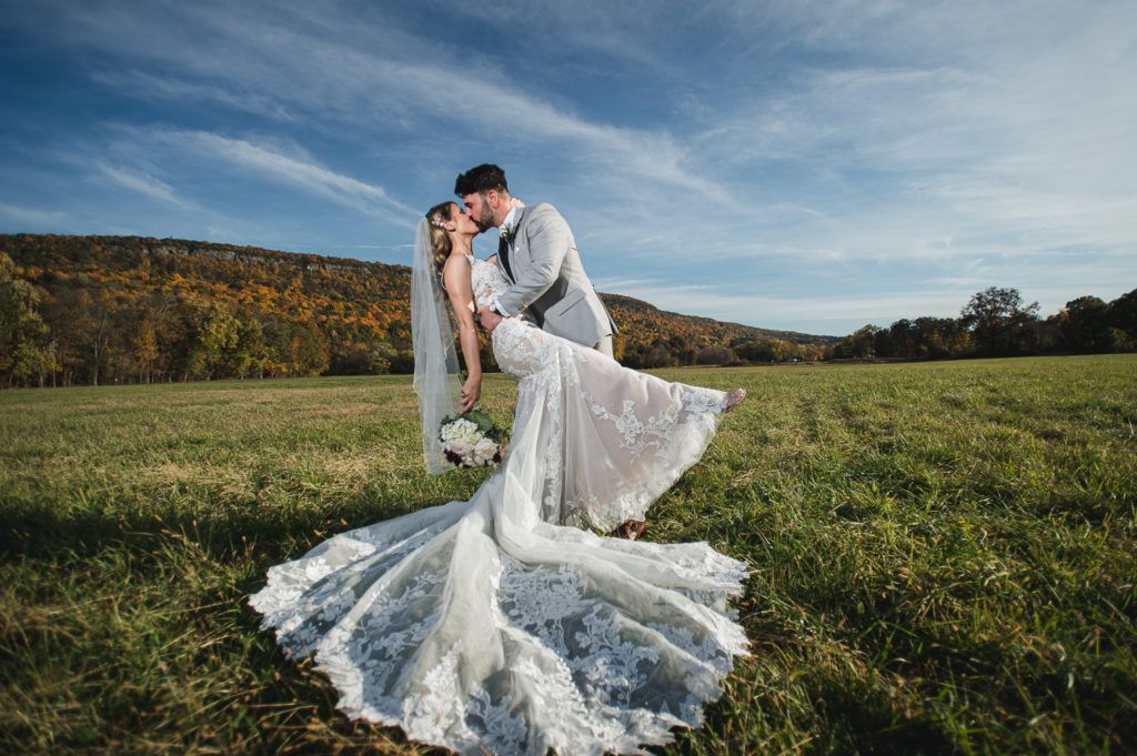 Couple kissing in a field, woman in a white lace wedding dress, man in a grey suit, blue sky.