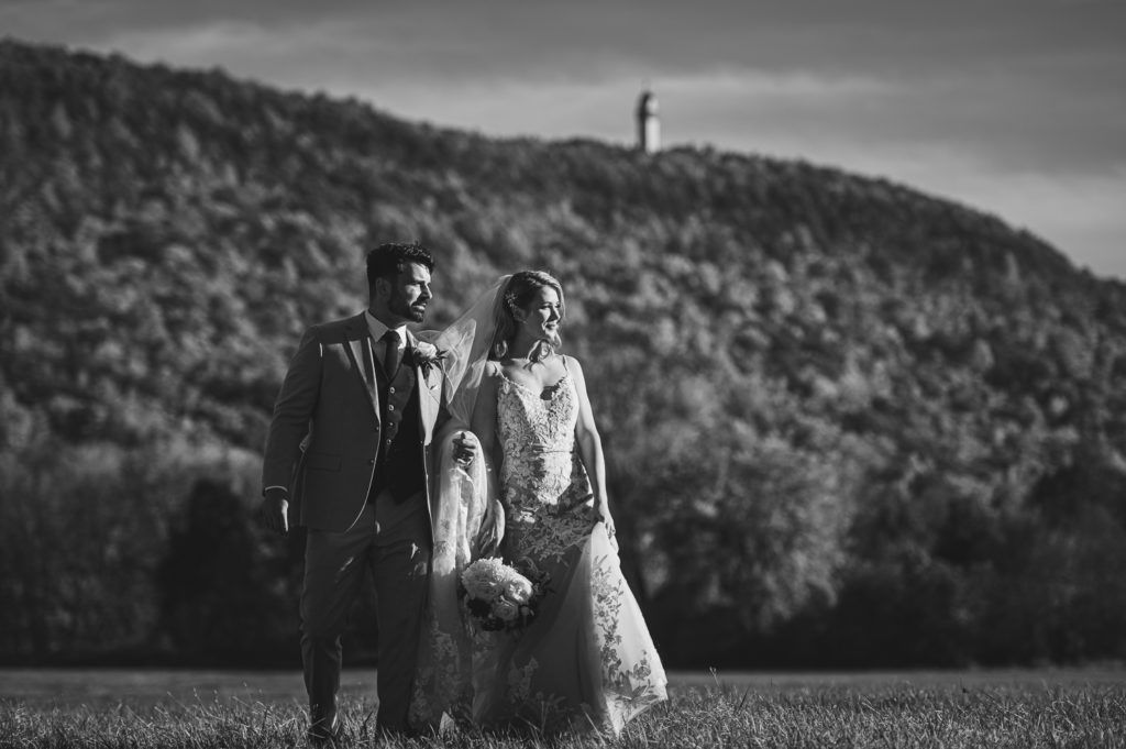 Couple in wedding attire walking in a field with a forested hill in the background. Black and white.