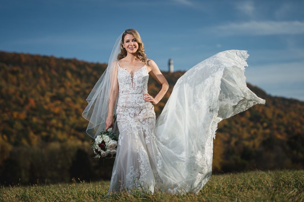 Bride in a white lace wedding dress, holding bouquet, veil flowing, posing in a field, mountain backdrop.