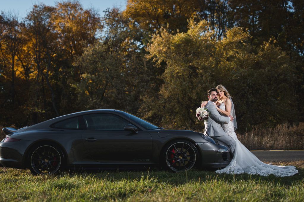 Newlyweds embrace next to a gray Porsche on a sunny road; autumn trees in the background.