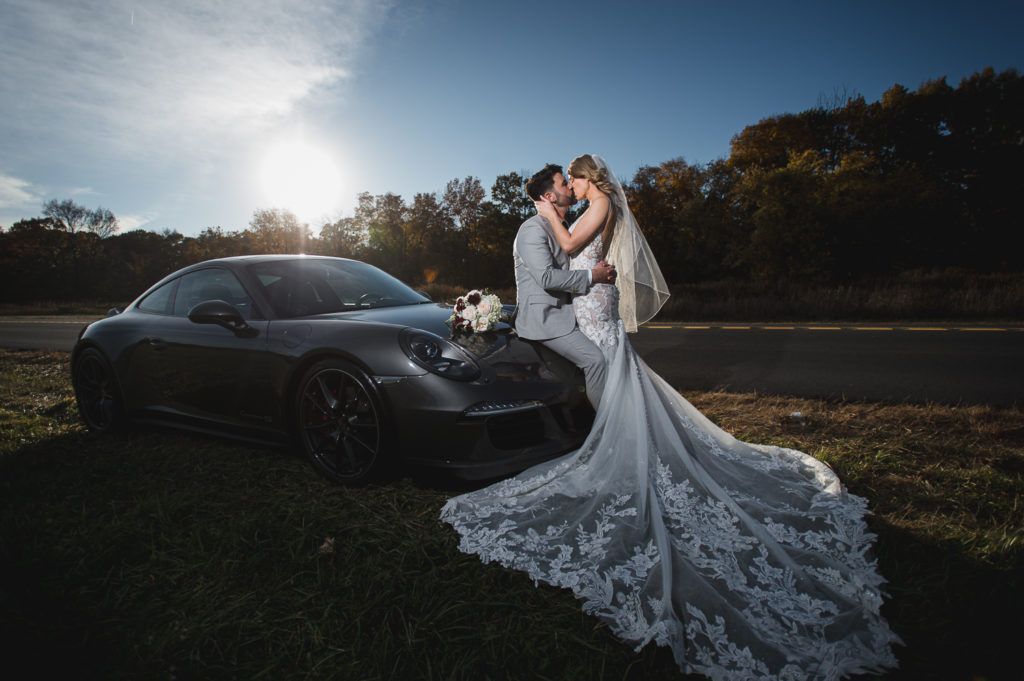 Newlyweds kiss next to a dark gray sports car. Bride's train spreads on hood; sunny outdoor setting.