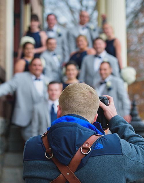 Photographer taking a group photo of a wedding party on steps; people in gray suits and dark dresses.