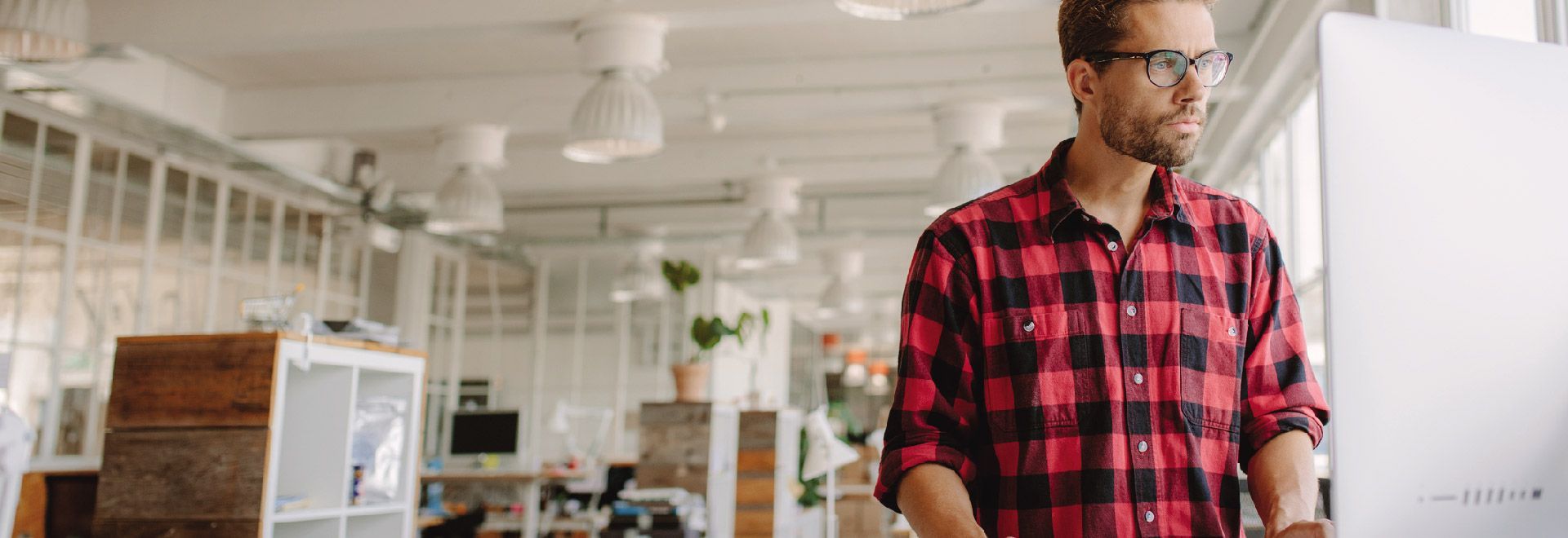 A man in a plaid shirt is standing in front of a computer in an office.
