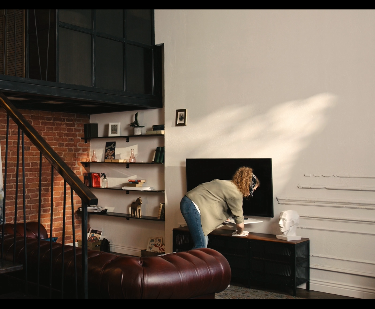 A woman in jeans and a shirt adjusts objects on a TV stand in a loft apartment with a brick wall and leather couch.
