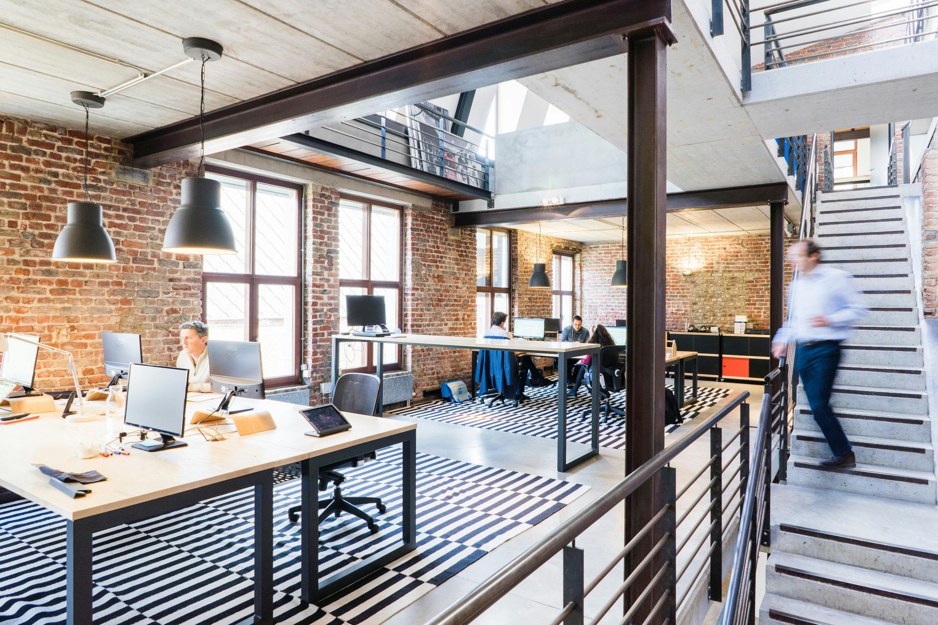 Modern office interior with brick walls, exposed beams, and multiple workstations; a person walks down stairs.