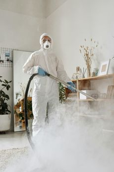 Person in hazmat suit spraying disinfectant in a room, creating a cloud of mist over a carpet.