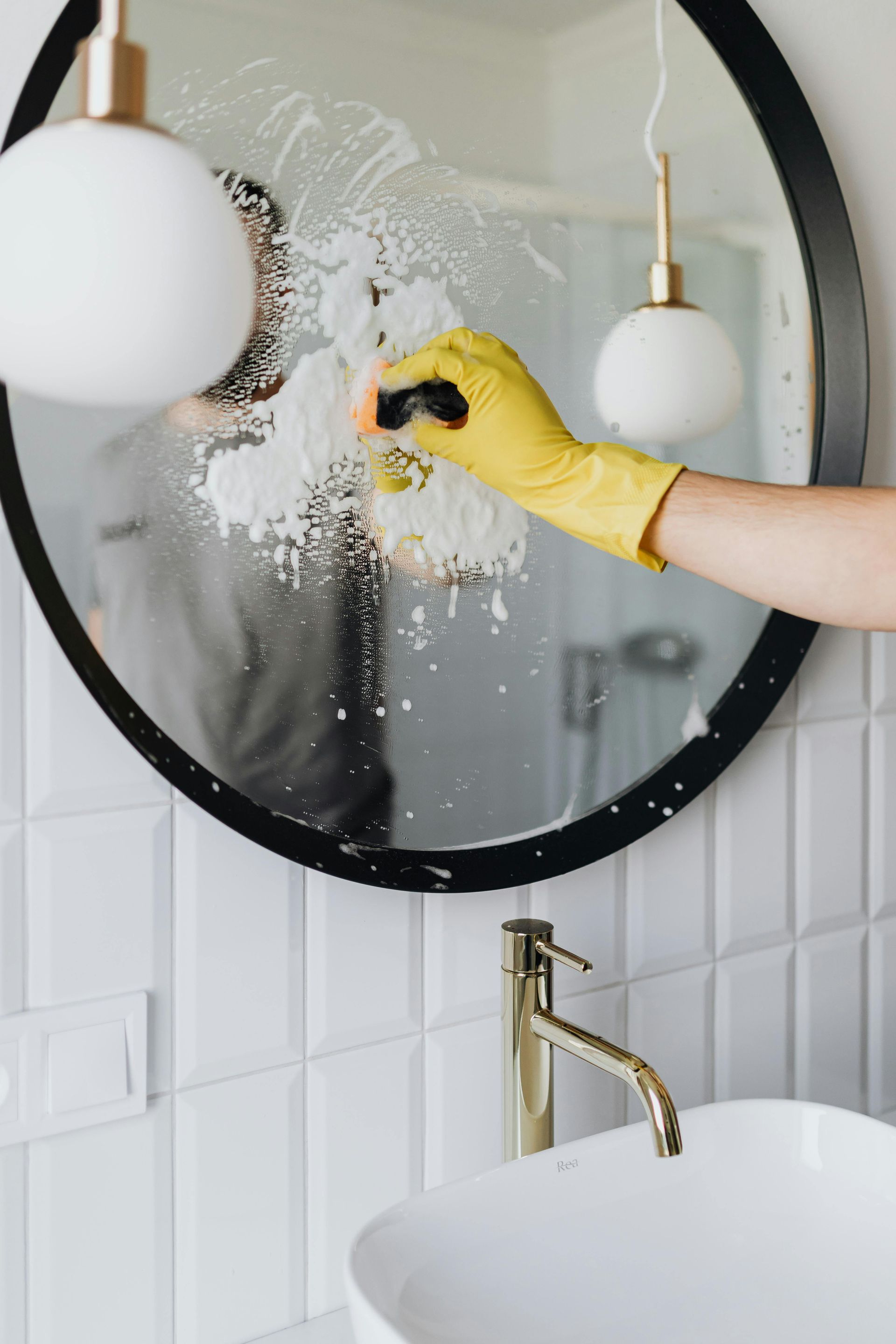Gloved hand spraying cleaner on a round bathroom mirror above a white sink and brass faucet