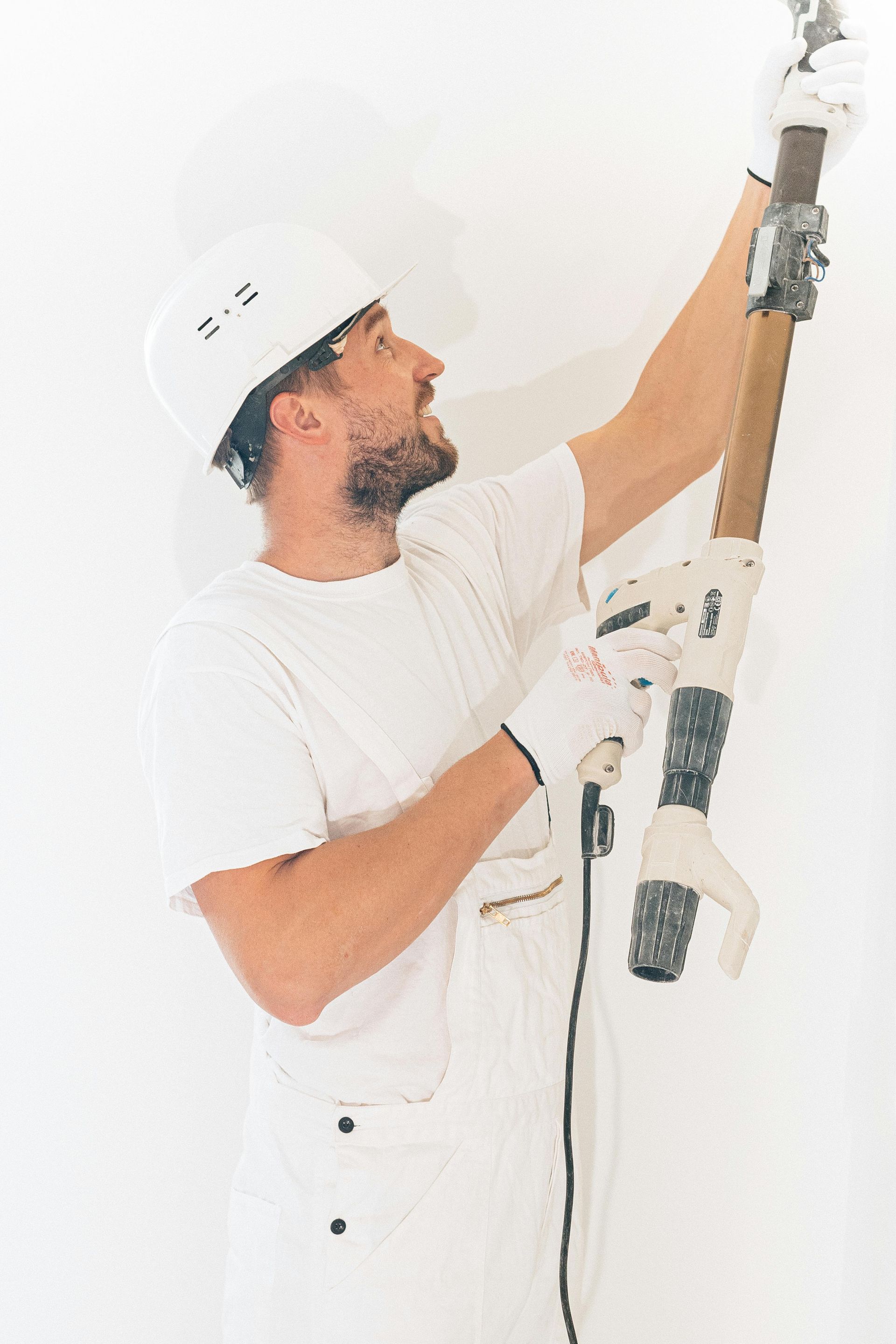 Man in white work clothes and hard hat, sanding a wall with a power sander, indoors.