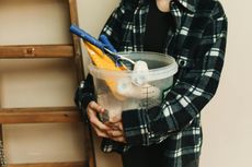 Person holding a clear paint bucket filled with tools, standing near a wooden ladder, wearing a black and white plaid shirt.