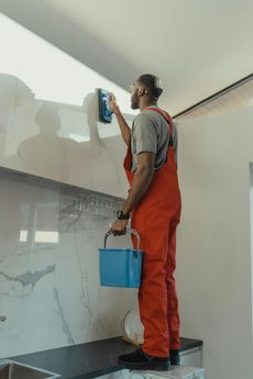 A person in red overalls and a grey shirt cleans a wall with a blue sponge while holding a bucket, standing on a black surface with a white wall background.