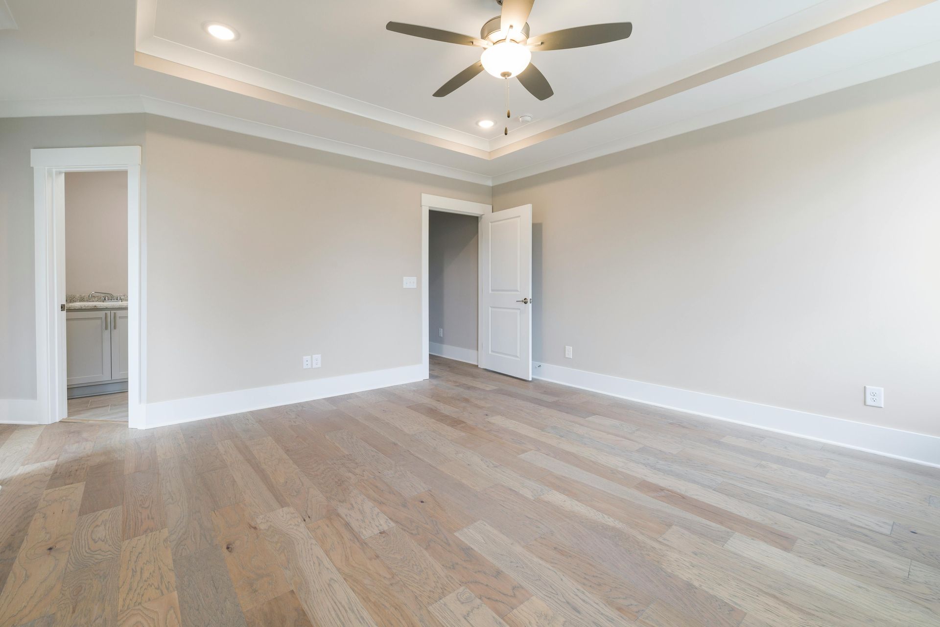 Empty modern bedroom with light gray walls, wood floors, tray ceiling, ceiling fan, and open closet door