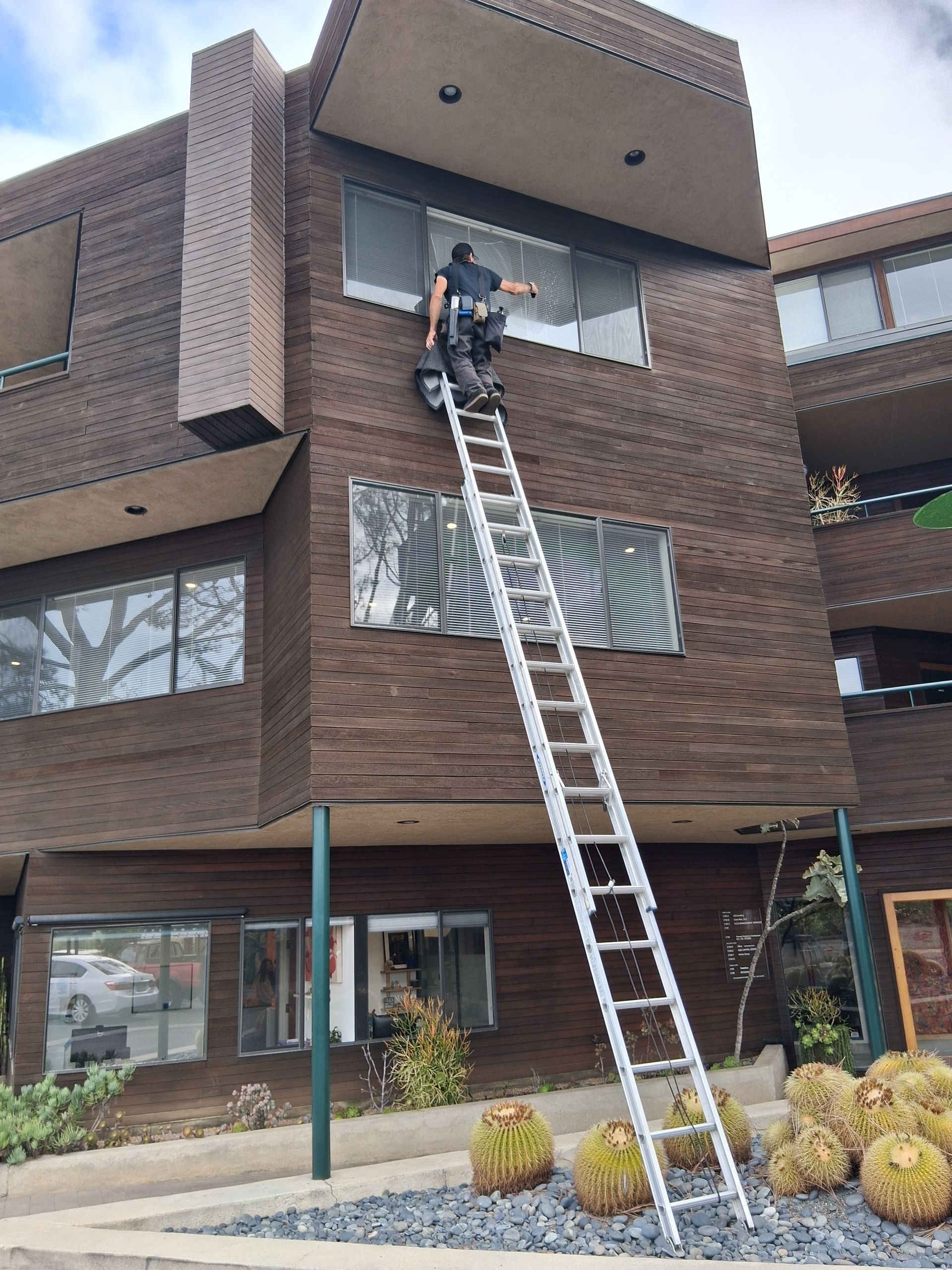 Person on ladder cleaning windows on a brown-sided building with balconies. Blue sky.