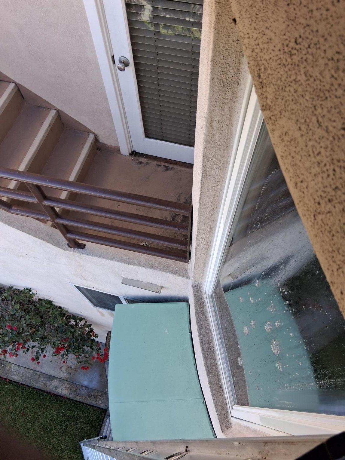 Exterior view: Building entrance with stairs, door, and a window. Beige building, brown railing, and green cushion visible.
