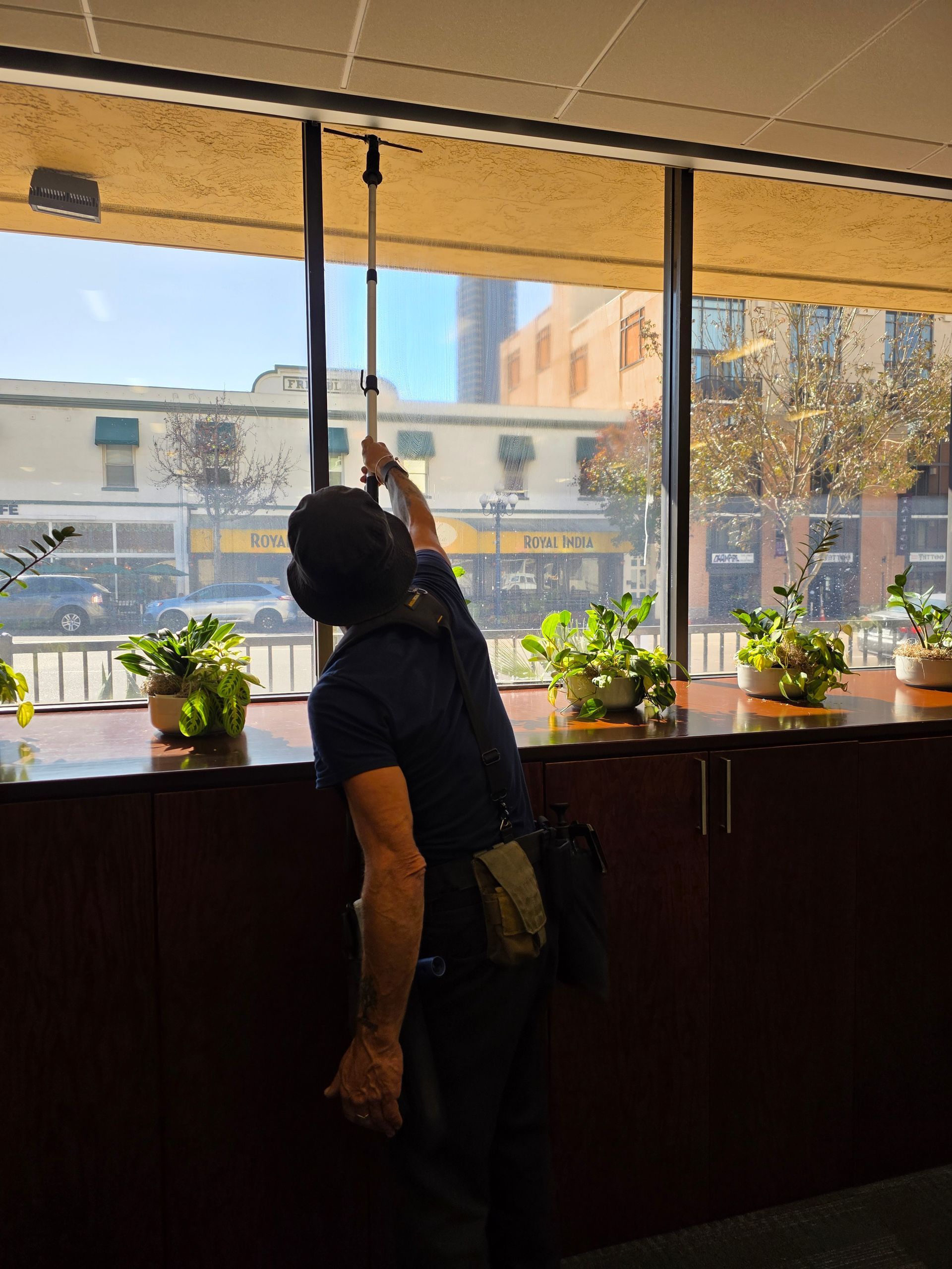 A person reaching up with a telescoping pole to adjust a window blind indoors, facing a sunny city view.