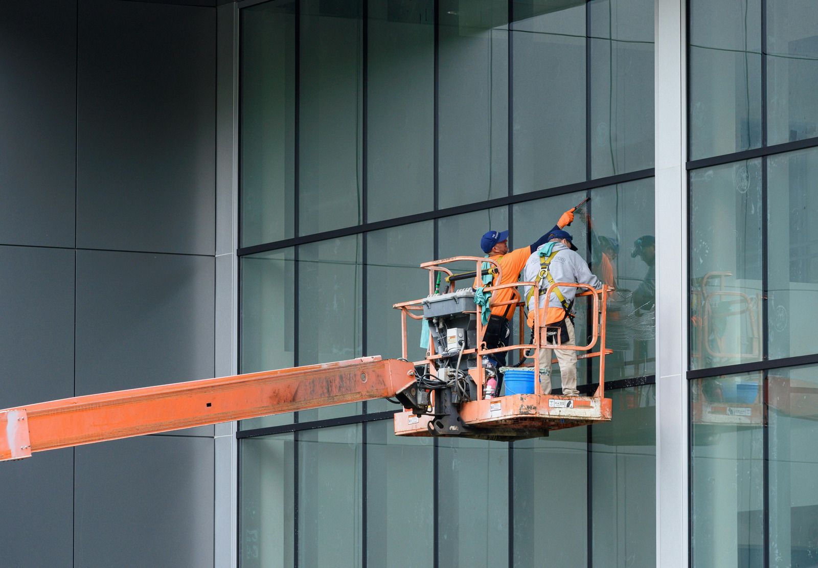 Two window washers in a lift cleaning a tall glass building, wearing safety harnesses and orange vests.