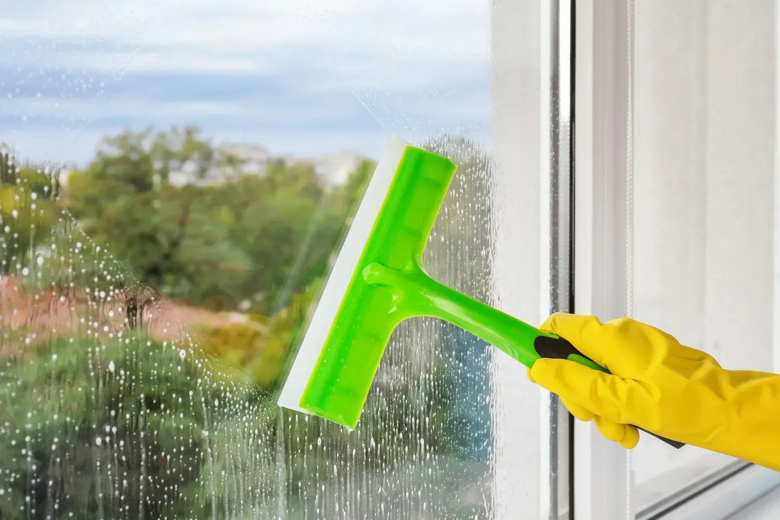 Hand in yellow glove using green squeegee to clean a window.