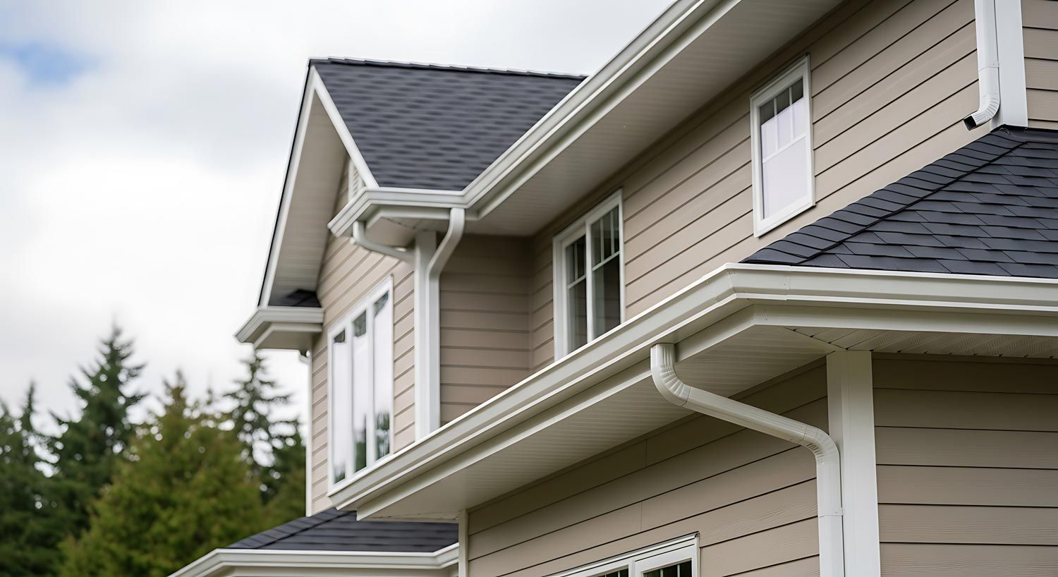 Close-up of a beige house exterior with white trim, gray shingle roof, and evergreen trees behind it