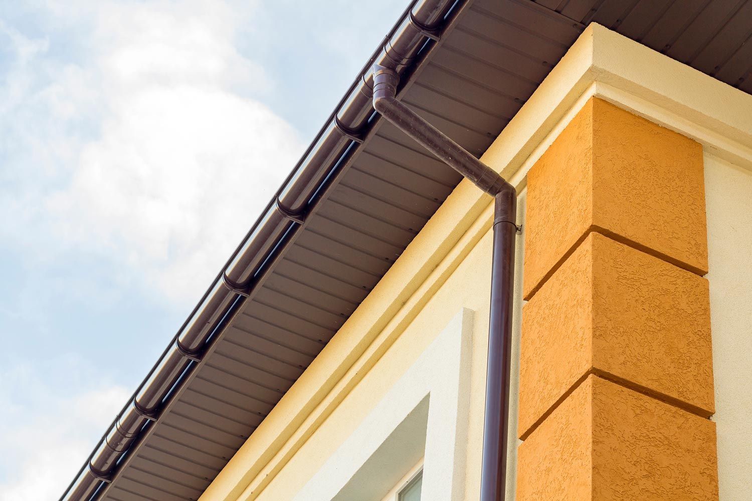 Corner of a house with orange stucco walls, white trim, and dark roof gutters against a cloudy sky