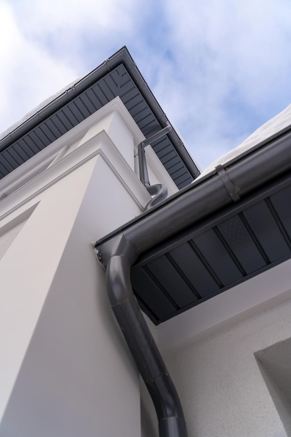 Upward view of a white house exterior with dark gray rooflines and a gutter against a cloudy sky
