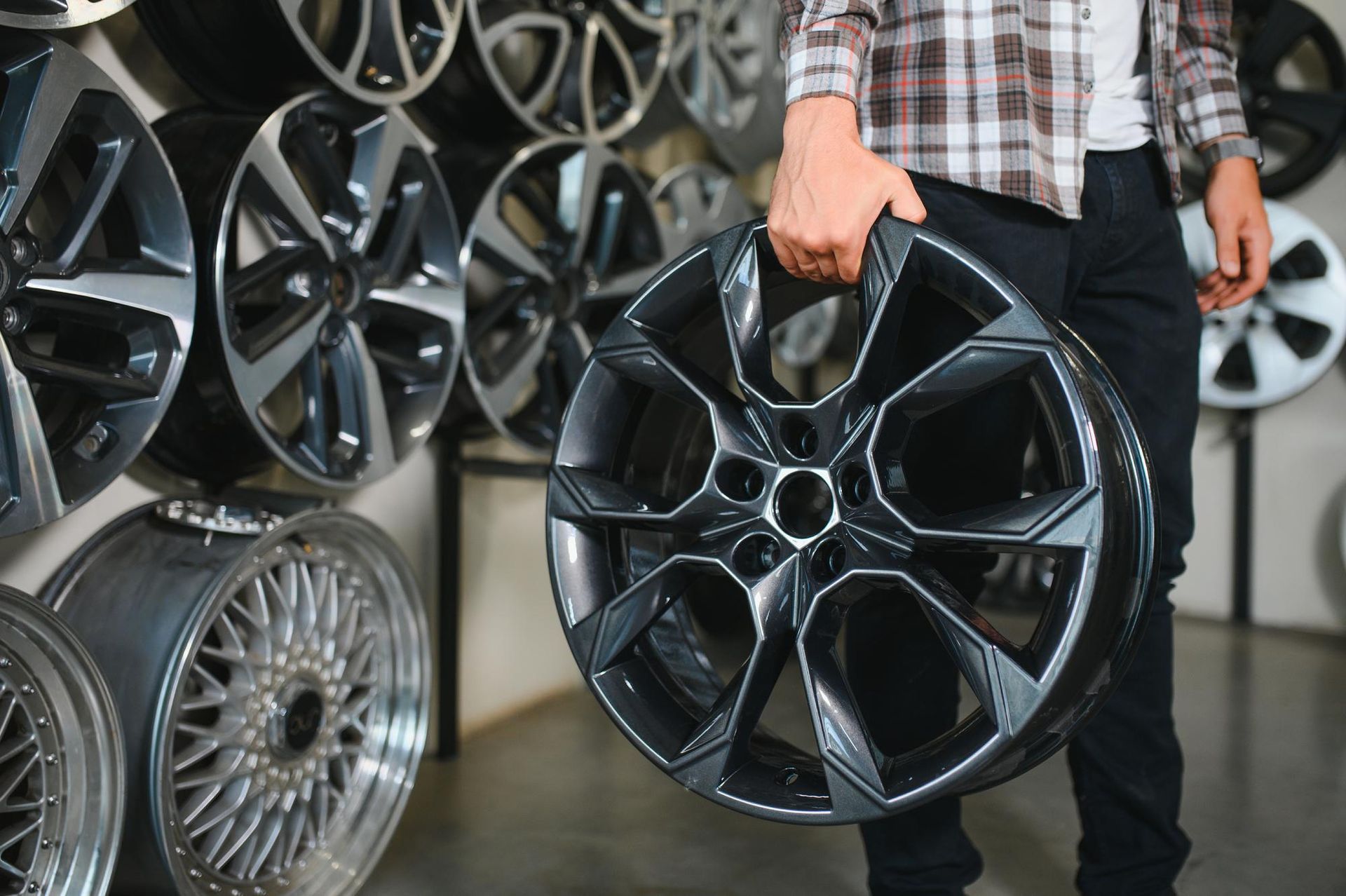 Man holding a black car rim in a shop, with a wall of rims in the background.