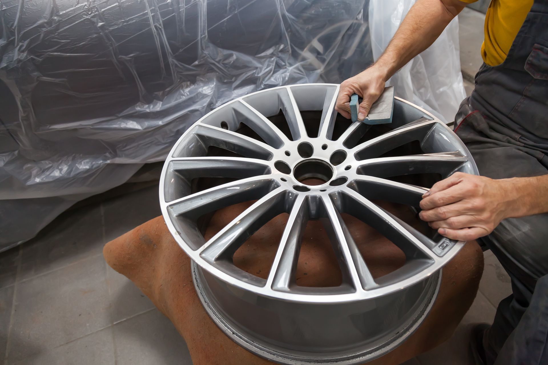 Person sanding a silver car wheel in a repair shop.